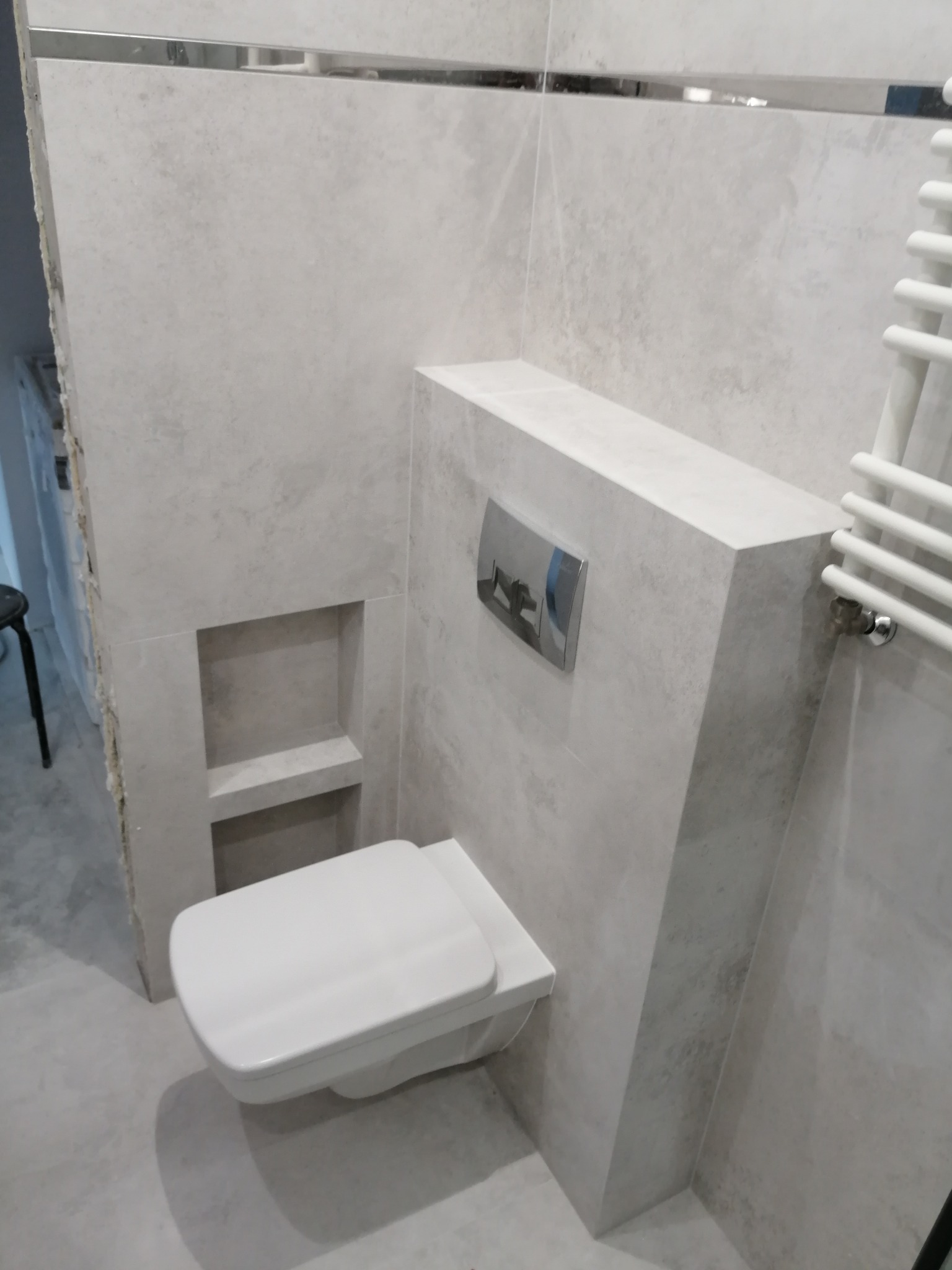 Ściana wyłożona jasnymi płytkami imitującymi kamień, z wbudowaną toaletą podtynkową, półką i grzejnikiem drabinkowym w łazience.