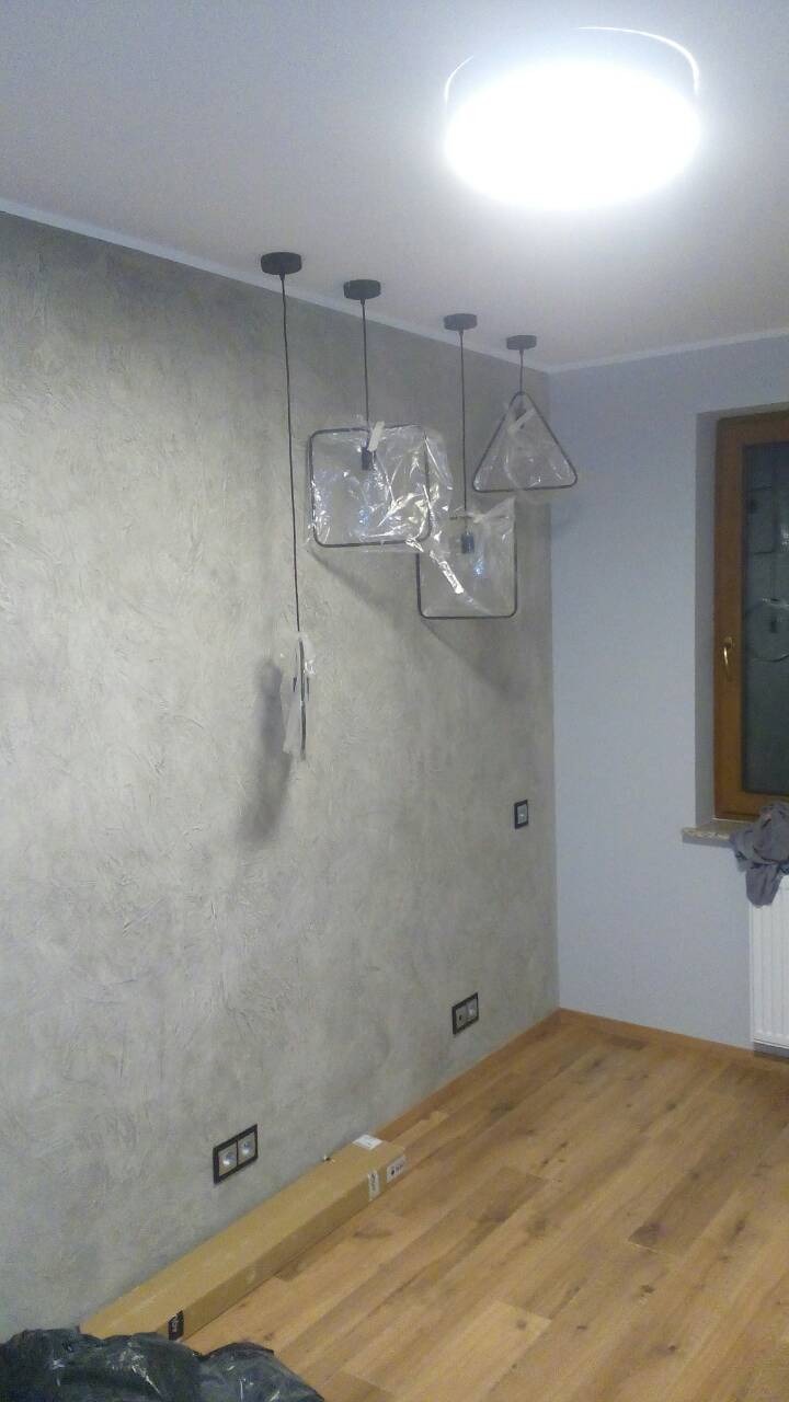 Pokój po remoncie z nową teksturą na ścianie, drewnianą podłogą i trzema nowoczesnymi lampami wiszącymi w plastikowych osłonach ochronnych.
