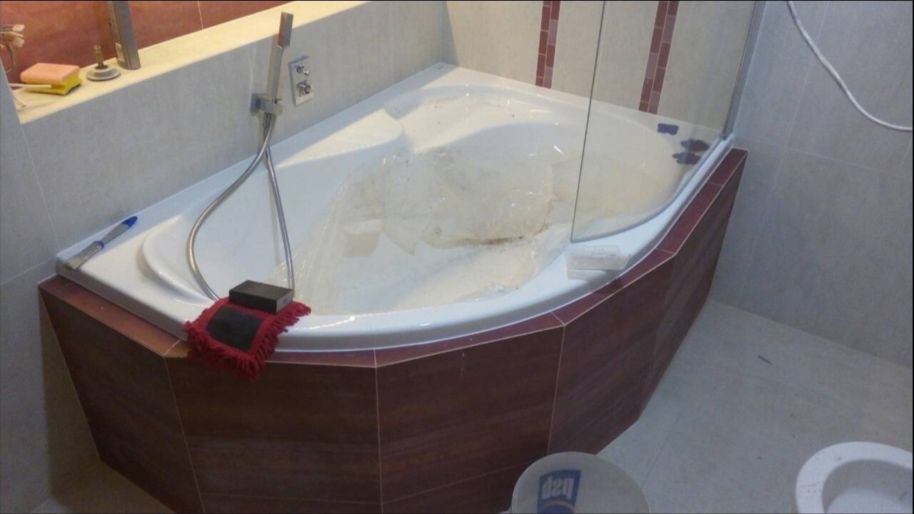 Narożna wanna z hydromasażem, zabezpieczona folią podczas prac wykończeniowych, widoczny fragment kabiny prysznicowej, akcesoria glazurnicze na brzegu wanny oraz fragment toalety w tle.
