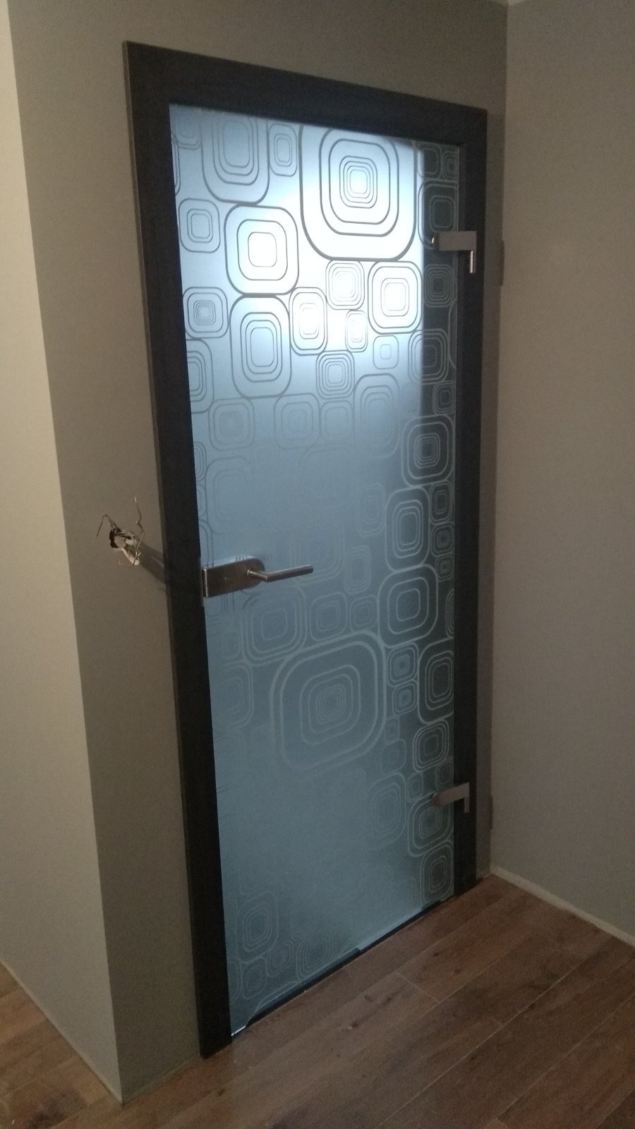 Szklane drzwi z geometrycznym, matowym wzorem kwadratów, zamontowane w ciemnej ramie, z widocznym metalowym uchwytem i zawiasami, osadzone w ścianie z widocznymi śladami po wcześniejszym montażu...