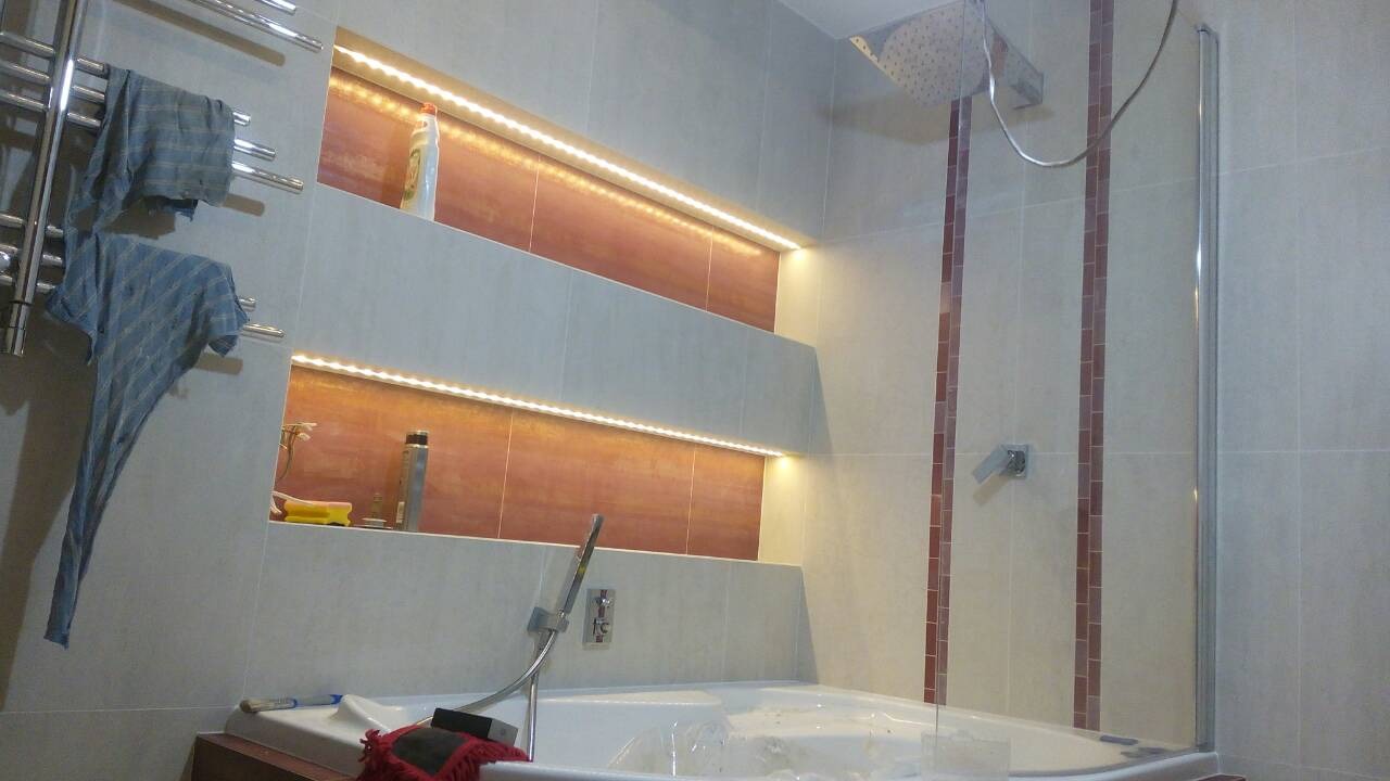 Nowoczesna łazienka z wanną z hydromasażem, podświetlanymi niszami w ścianie wyłożonej płytkami w odcieniach beżu i brązu, oraz szklanym panelem prysznicowym.