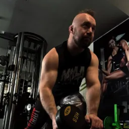 Mężczyzna z brodą, w sportowym stroju, ćwiczy z 16kg hantlą w siłowni. Na ścianie grafika kobiety w stroju sportowym. Ujęcie z dołu, podkreślające muskulaturę.