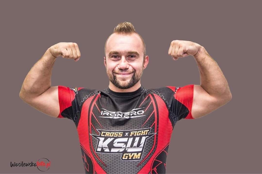 Uśmiechnięty mężczyzna z irokezem prezentuje umięśnione bicepsy, ubrany w koszulkę z logo KSW Cross Fight Gym na szarym tle.