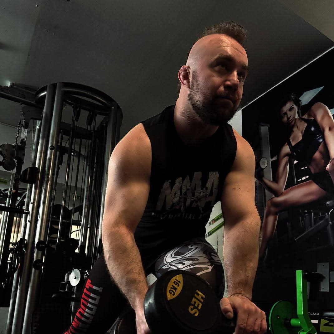 Mężczyzna z brodą, w sportowym stroju, ćwiczy z 16kg hantlą w siłowni. Na ścianie grafika kobiety w stroju sportowym. Ujęcie z dołu, podkreślające muskulaturę.