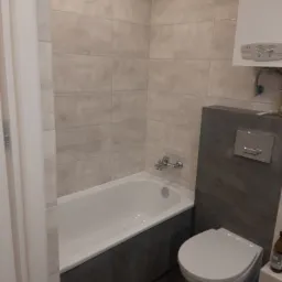 Wykończona łazienka z wanną obudowaną szarymi płytami, wiszącą toaletą, beżowymi płytkami na ścianach i grzejnikiem gazowym nad toaletą.