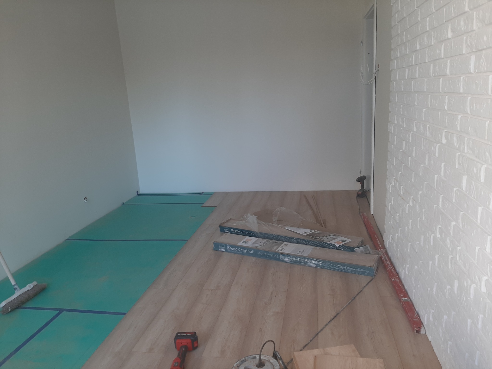 Wnętrze w trakcie prac: częściowo ułożone panele podłogowe w odcieniach beżu, widoczne paczki paneli Krono Original, podkład pod panele w kolorze zielonym, narzędzia (poziomica, wiertarka, młotek...