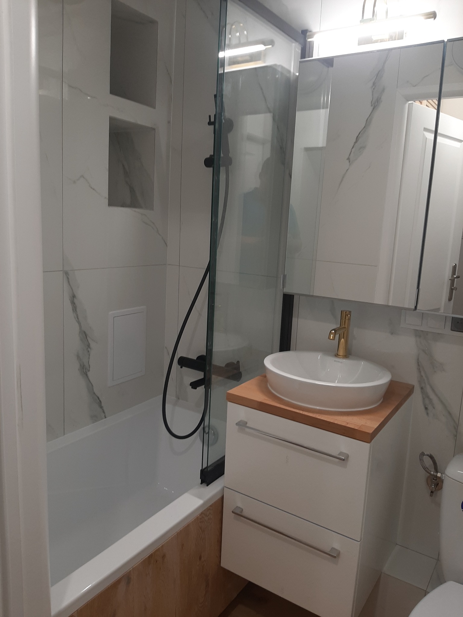 Nowoczesna łazienka z wanną obudowaną drewnem, kabiną prysznicową z czarną armaturą, umywalką nablatową na szafce z drewnianym blatem i marmurowymi płytkami na ścianach.