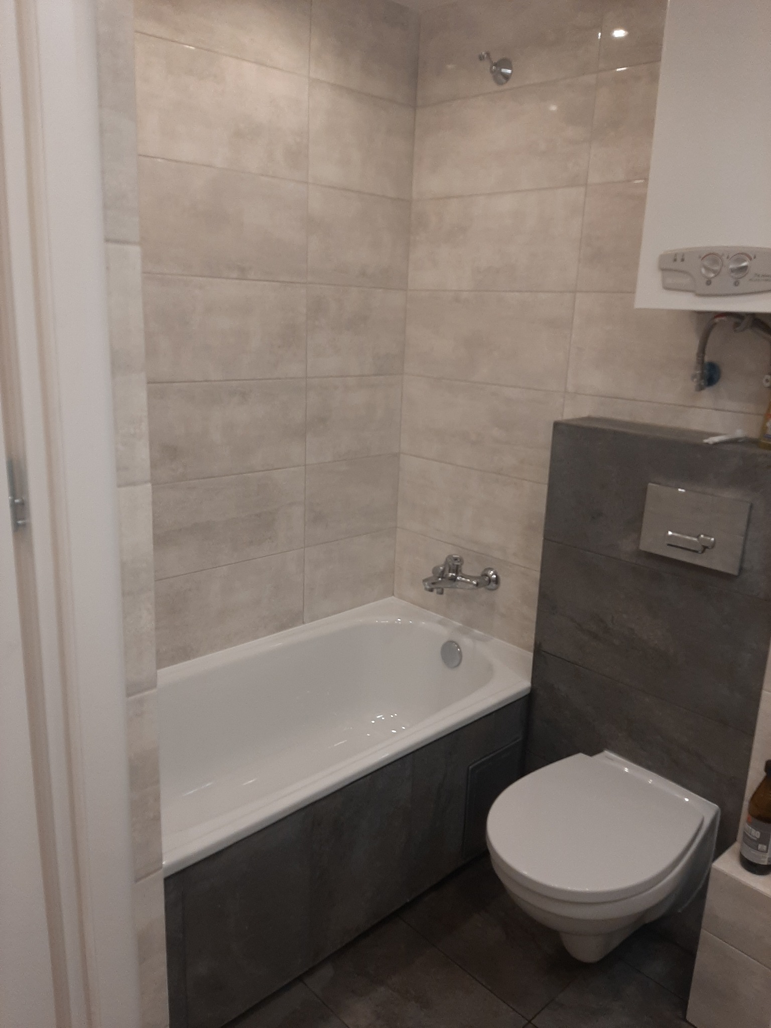 Wykończona łazienka z wanną obudowaną szarymi płytami, wiszącą toaletą, beżowymi płytkami na ścianach i grzejnikiem gazowym nad toaletą.