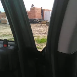 Widok z wnętrza samochodu na plac budowy z murarzami wznoszącymi ściany z czerwonej cegły, taczka w tle.