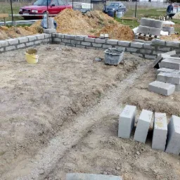 Wznoszenie fundamentów domu z bloczków betonowych na działce, widoczne wykopane rowy, zaprawa murarska, wiadro i samochody w tle.