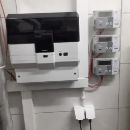 Nowoczesna rozdzielnica elektryczna z trzema licznikami FAUN na białej, kafelkowanej ścianie, widoczne kable i zasilanie.