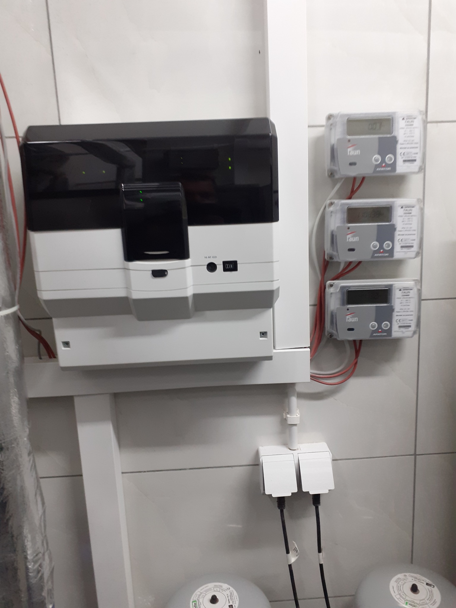 Nowoczesna rozdzielnica elektryczna z trzema licznikami FAUN na białej, kafelkowanej ścianie, widoczne kable i zasilanie.