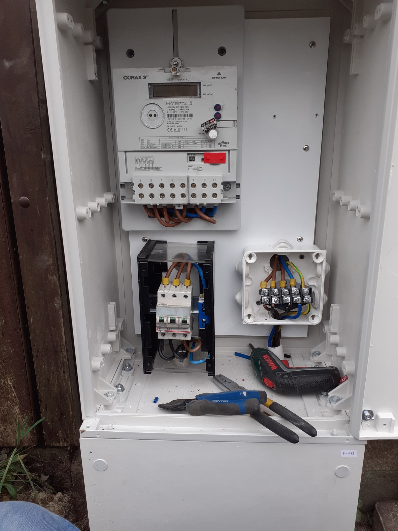 Otwarta skrzynka elektryczna z zamontowanym licznikiem Corax 3' i zabezpieczeniami, widoczne narzędzia: kombinerki Dexter i wkrętarka Bosch, okablowanie w trakcie podłączania.