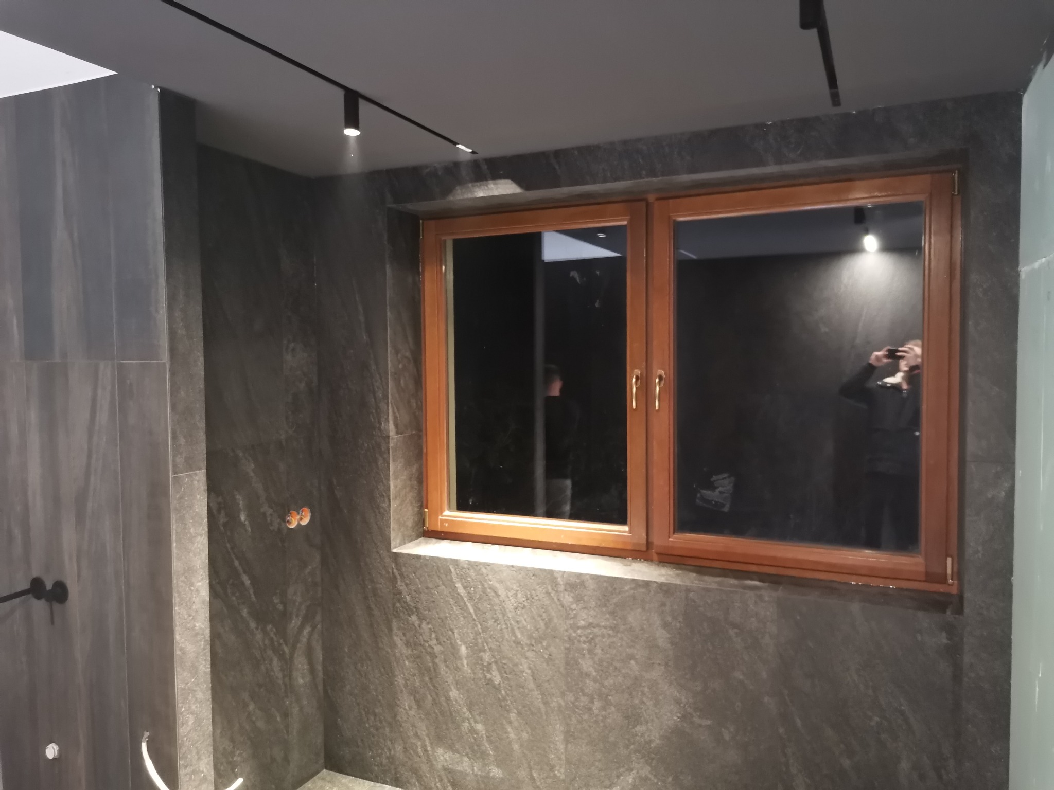 Nowoczesna łazienka z ciemnoszarą kamienną okładziną ścian, drewnianą ramą okienną i reflektorami sufitowymi na szynie; w odbiciu okna widoczna osoba robiąca zdjęcie.