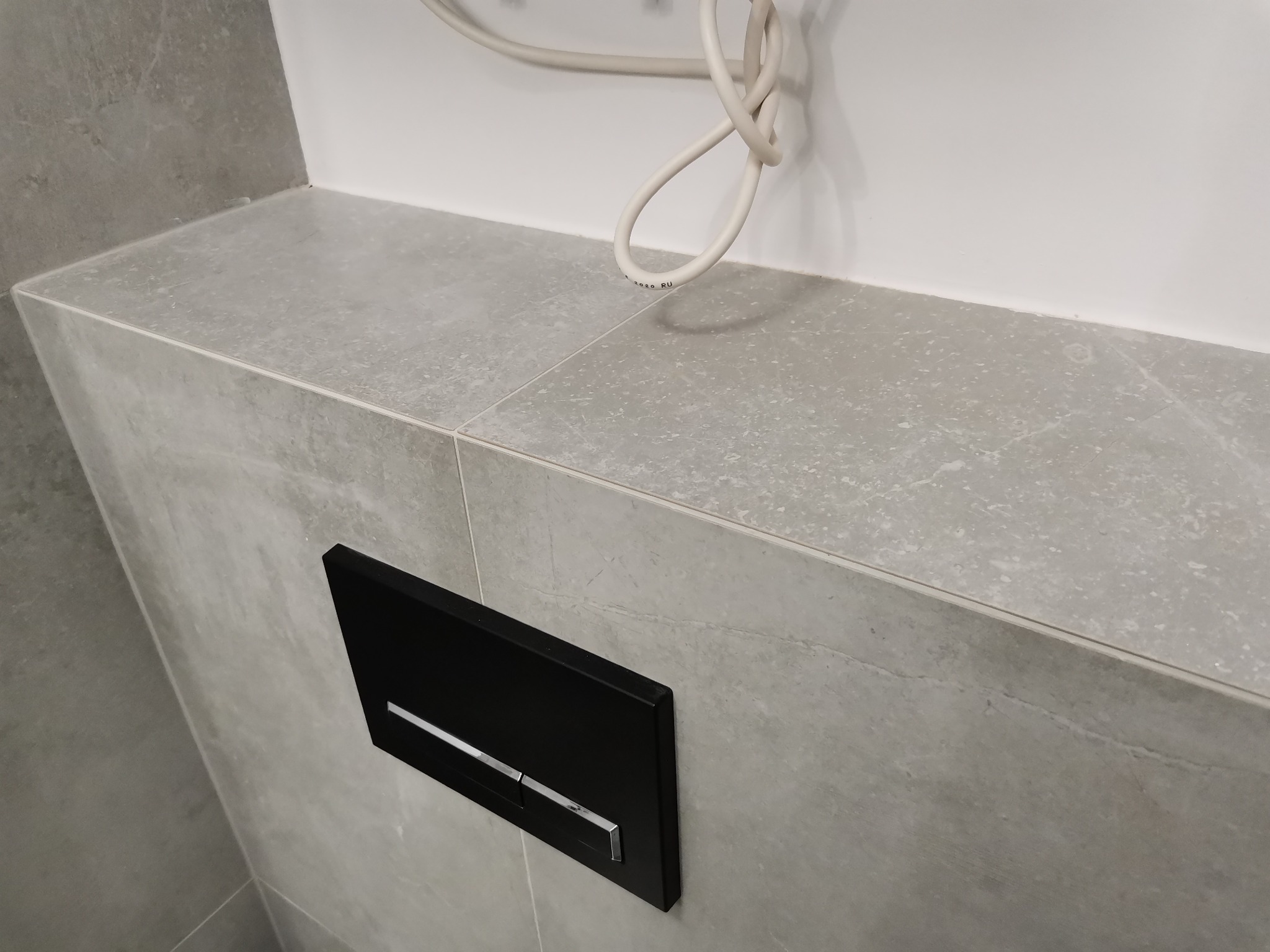 Szare płytki ceramiczne na ścianie z czarnym przyciskiem spłuczki WC z chromowanym paskiem, widoczne przewody instalacyjne.
