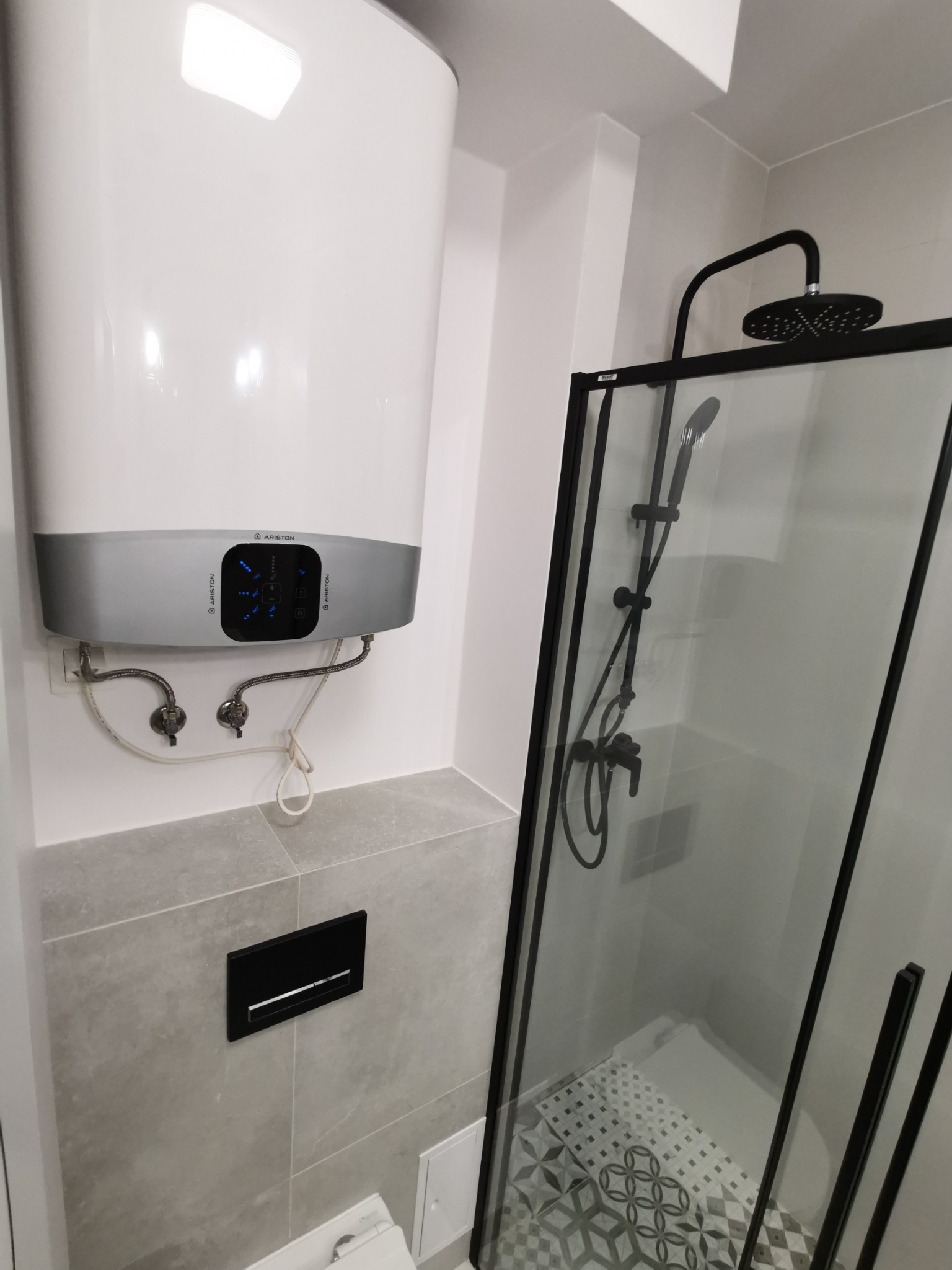 Biały, wiszący bojler Ariston z szarym panelem sterowania i podłączeniami hydraulicznymi, obok kabina prysznicowa z czarną armaturą i wzorzystymi płytkami podłogowymi w łazience wykończonej szarymi...