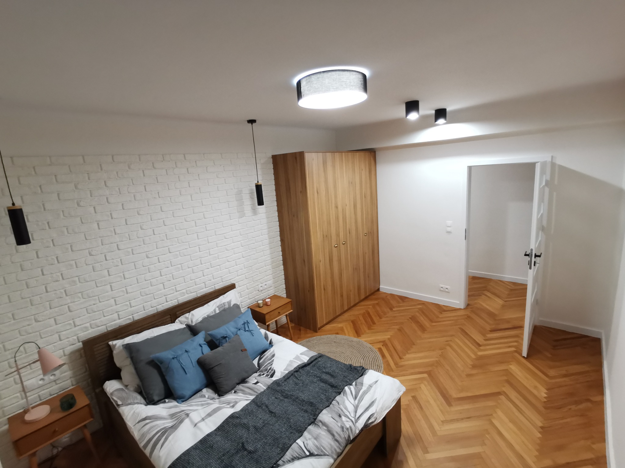 Jasna sypialnia z łóżkiem z drewnianą ramą, białą ceglaną ścianą, parkietem w jodełkę i drewnianą szafą na tle otwartych drzwi.