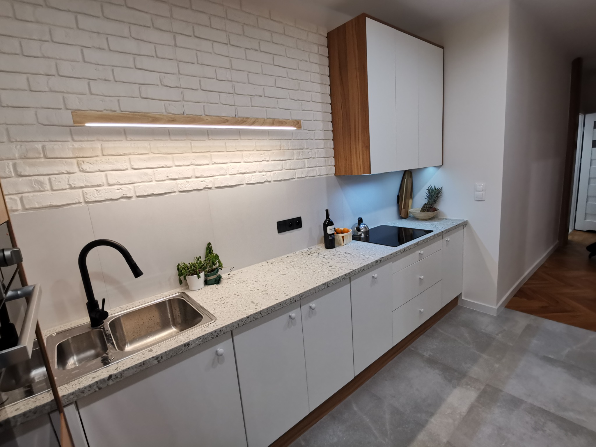 Nowoczesna kuchnia z białymi szafkami, blatem z lastryko i ścianą z białej cegły, oświetlona podłużną lampą LED. Na blacie widoczne akcesoria kuchenne i dekoracje.