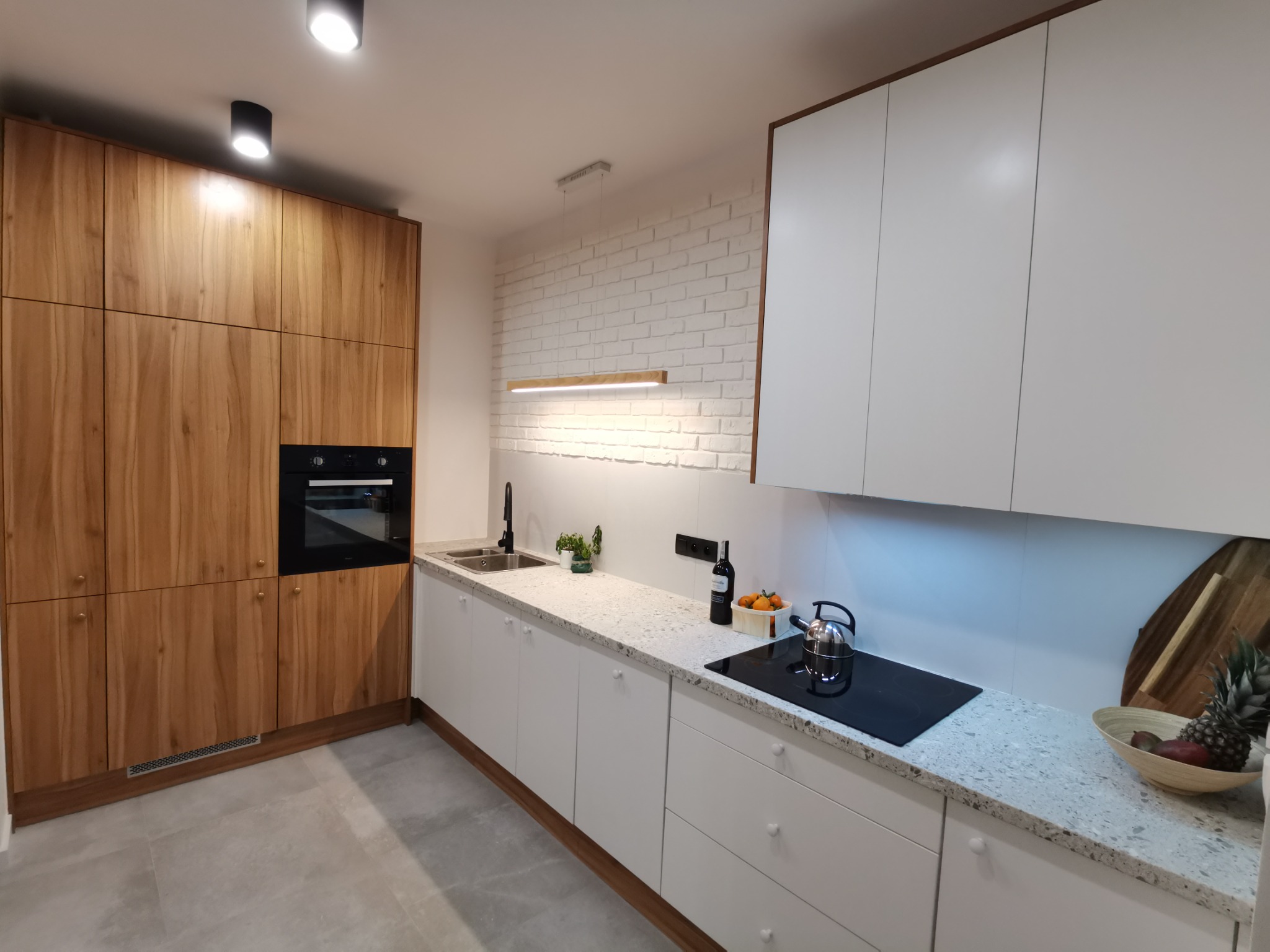 Nowoczesna kuchnia z drewnianymi i białymi szafkami, blatem z jasnego lastryko, czarną armaturą i płytą indukcyjną, oświetlona lampą wiszącą i spotami.