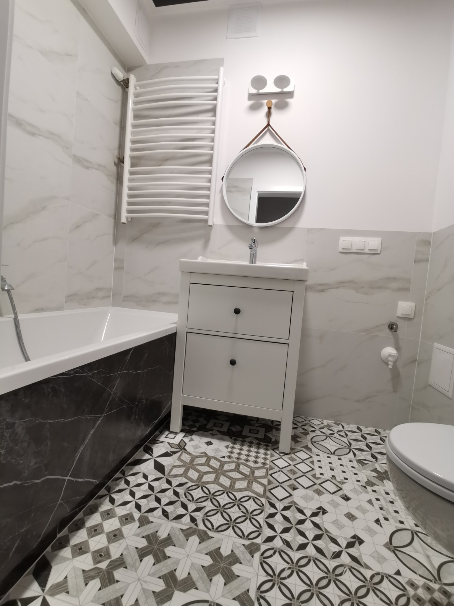 Nowoczesna łazienka z wanną obudowaną czarnym marmurem, białą szafką z umywalką, okrągłym lustrem na pasku i wzorzystą, patchworkową podłogą.