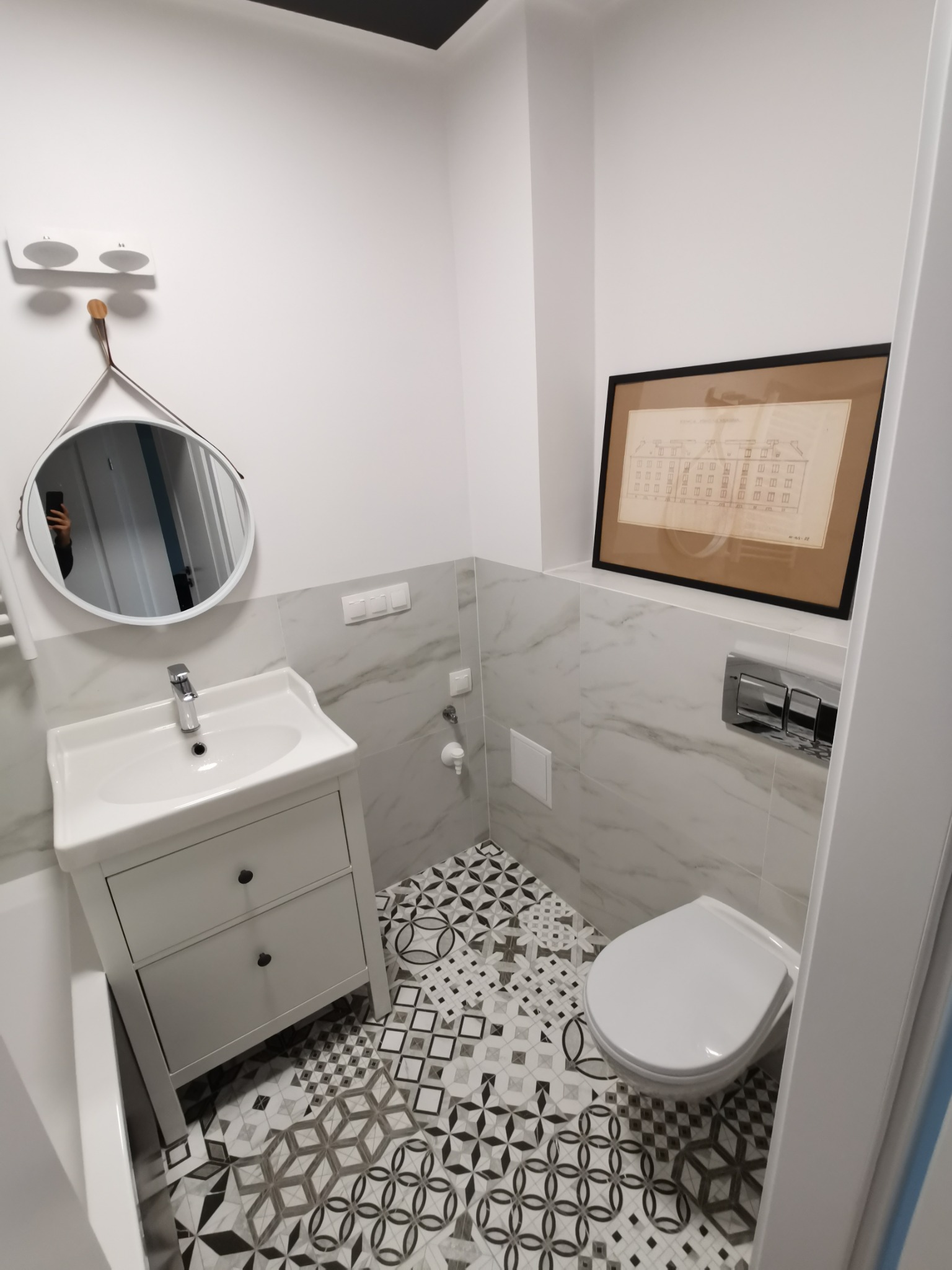 Nowoczesna łazienka z białą umywalką z szafką, okrągłym lustrem, toaletą, marmurowymi płytkami do połowy ściany i wzorzystą czarno-białą podłogą. Na ścianie wisi oprawiony plan architektoniczny.