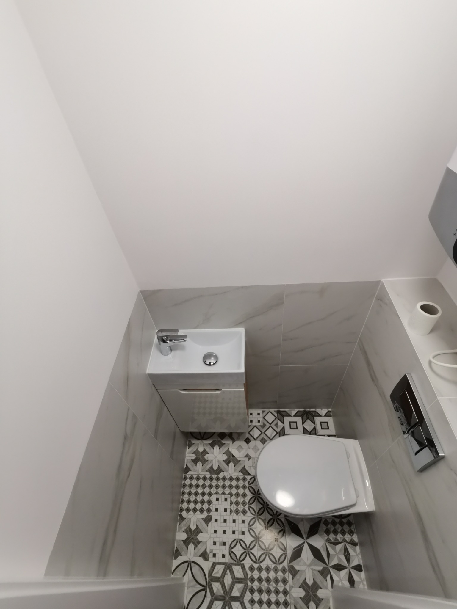 Niewielka łazienka w odcieniach szarości i bieli, z wzorzystą podłogą, wiszącą toaletą i umywalką z szafką, sfotografowana z góry.