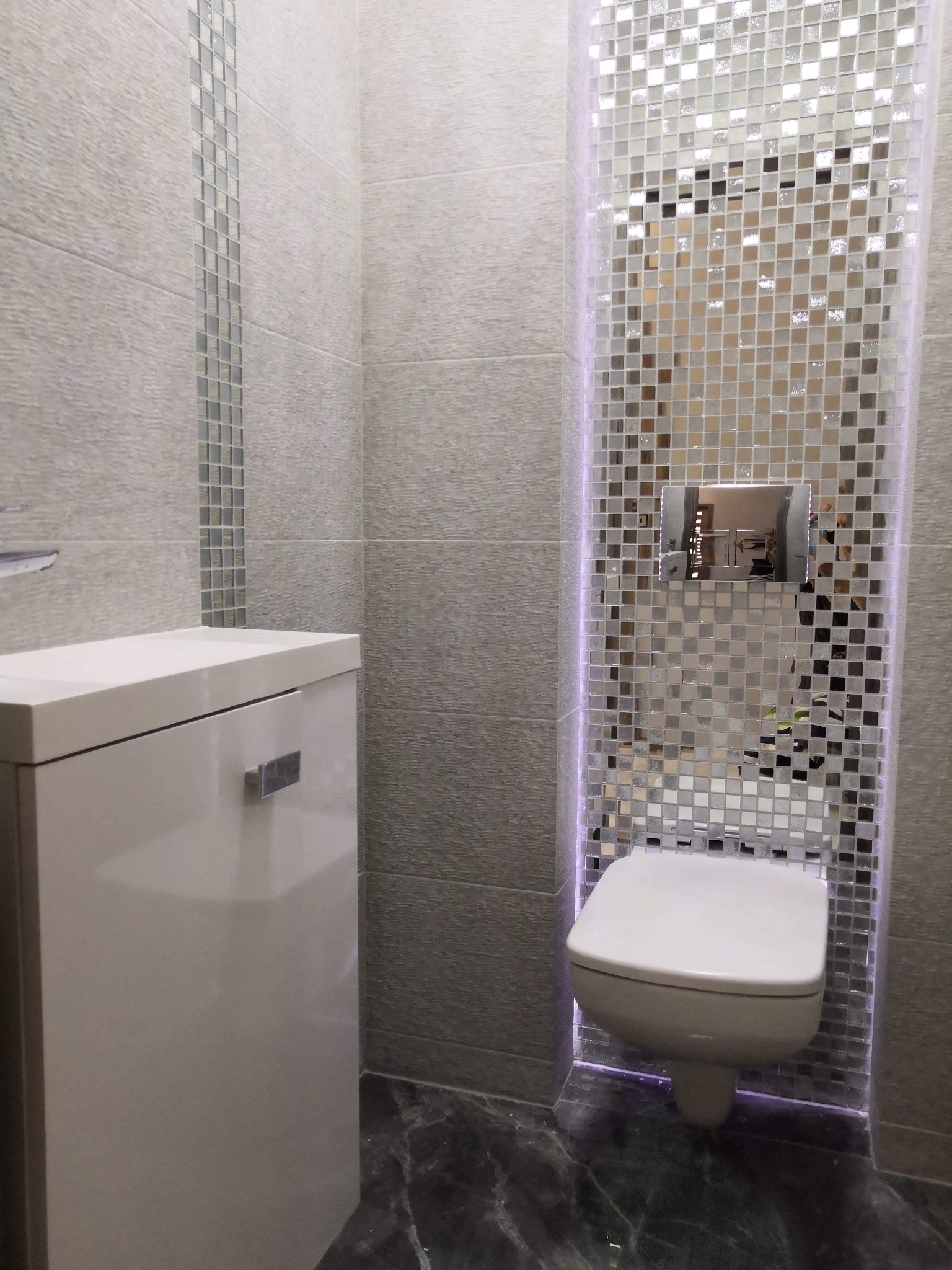 Nowoczesna toaleta podwieszana otoczona mozaiką z podświetleniem LED, obok zabudowa sanitarna i płytki ścienne z dekoracyjnym paskiem mozaiki, podłoga z ciemnego marmuru.
