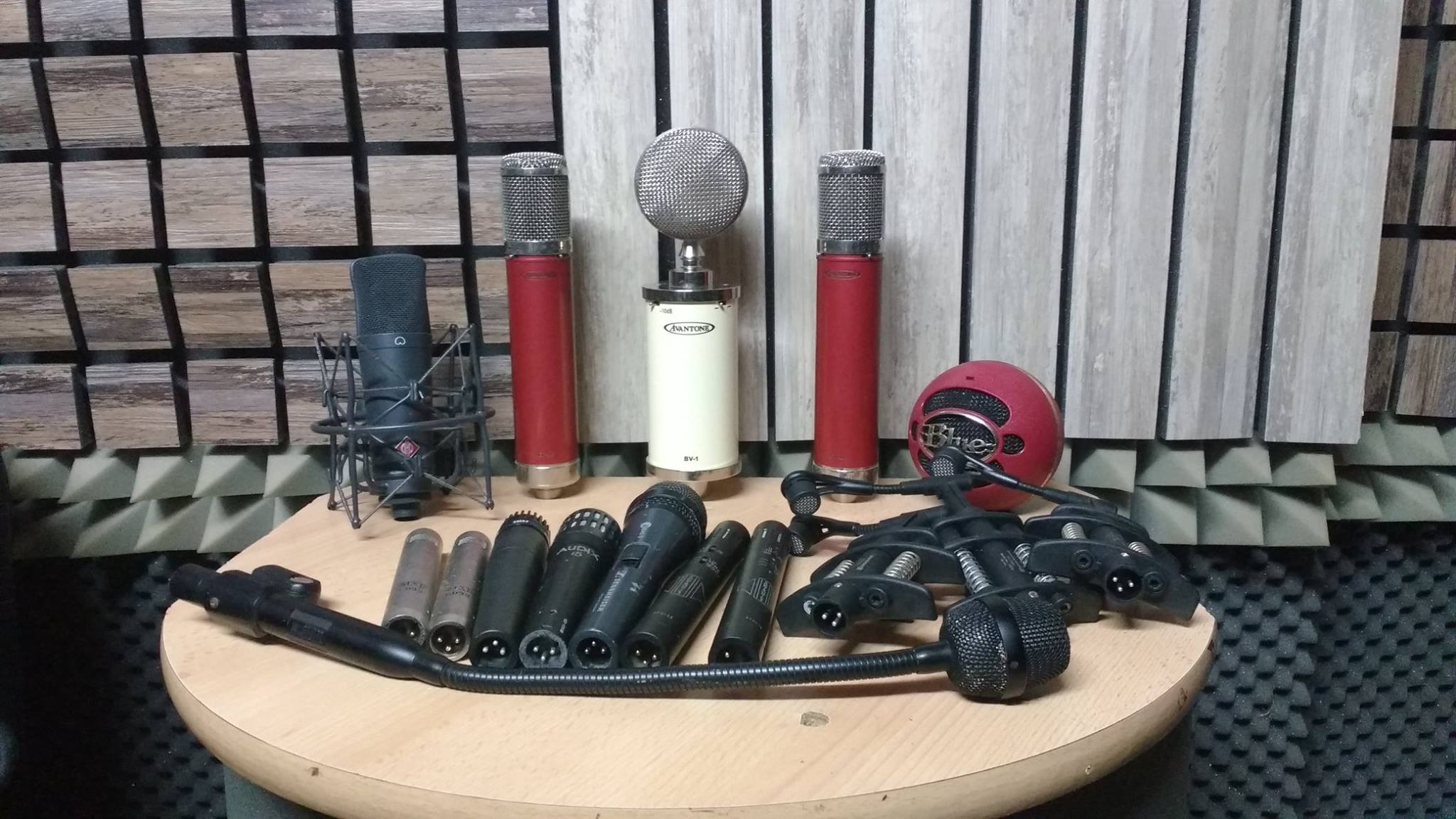 Kolekcja mikrofonów różnych typów i marek, w tym pojemnościowe i dynamiczne, ustawionych na drewnianym stole w pomieszczeniu z panelami akustycznymi na ścianach.