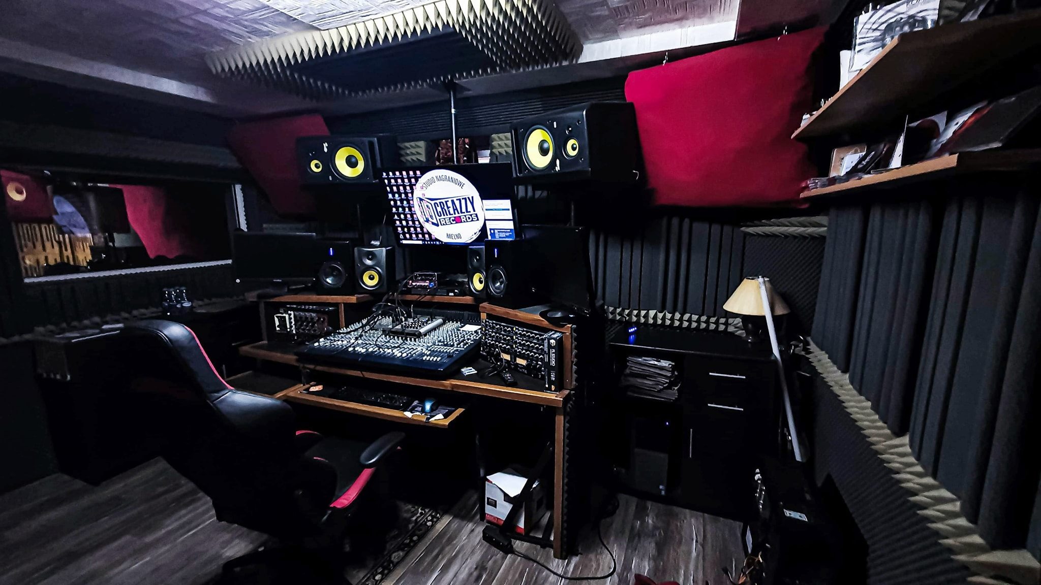 Profesjonalne pomieszczenie z panelami akustycznymi, mikserem dźwięku, monitorami studyjnymi i komputerem z logo 'Creazzy Records'.