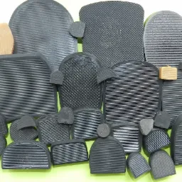 Zestaw różnorodnych, gumowych fleczków do butów w odcieniach szarości i beżu, z różnymi fakturami powierzchni, ułożonych na jasnozielonym tle.