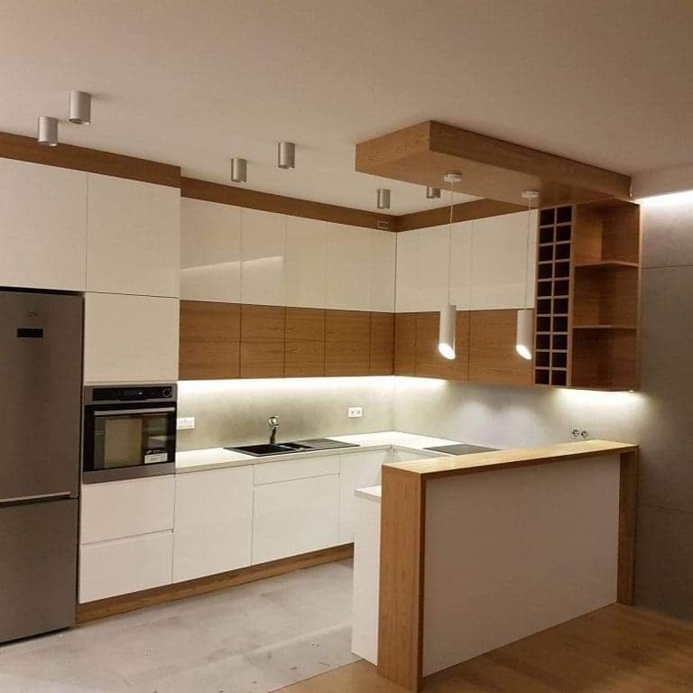 Nowoczesna kuchnia z białymi, lakierowanymi frontami i drewnianymi akcentami, podświetleniem LED pod szafkami wiszącymi, wyspą z blatem z drewna i zabudowaną półką na wino.