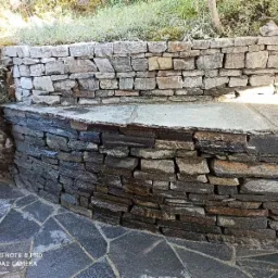 Kamienna ściana oporowa z nieregularnych płaskich kamieni, tworząca schody lub taras, z podłogą wyłożoną płytami z ciemnego kamienia łupkowego.