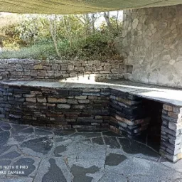 Kamienna zabudowa grilla zewnętrznego z blatem i wnęką na drewno, wykonana z ciemnego kamienia łupanego, na tle kamiennej ściany i posadzki z nieregularnych płyt kamiennych.