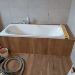 Wanna obudowana drewnopodobną okładziną, poziomica na krawędzi, widoczne narzędzia i materiały budowlane, płytki na ścianach w trakcie układania.