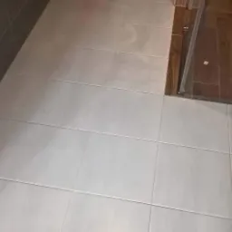 Jasnoszare, kwadratowe płytki podłogowe w łazience, z widoczną toaletą podwieszaną i fragmentem kabiny prysznicowej z drewnianym wykończeniem.