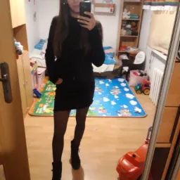 Kobieta robi selfie w lustrze, ubrana w czarną sukienkę i botki, w tle pokój dziecięcy z zabawkami i dywanem.