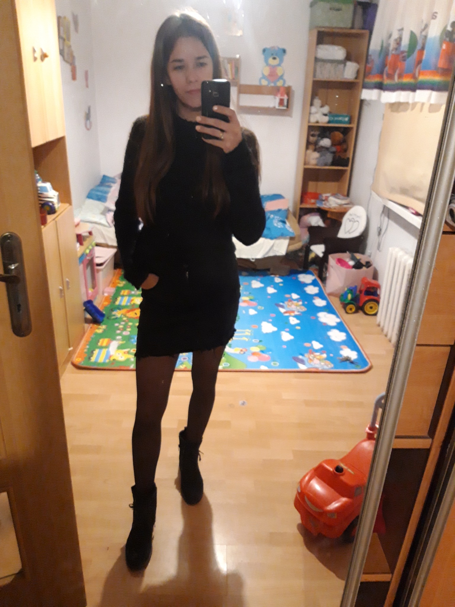 Kobieta robi selfie w lustrze, ubrana w czarną sukienkę i botki, w tle pokój dziecięcy z zabawkami i dywanem.
