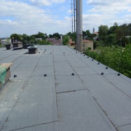 P.H.U. Dr Volt Zbigniew Frejman - Szary, płaski dach pokryty papą z elementami instalacji odgromowej i kominami wentylacyjnymi. W tle zabudowa miejska i błękitne niebo z chmurami.