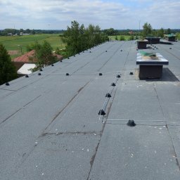 P.H.U. Dr Volt Zbigniew Frejman - Szary płaski dach pokryty papą z systemem odgromowym, widok z góry na tle zieleni i błękitnego nieba z chmurami. Widoczne kominy wentylacyjne.