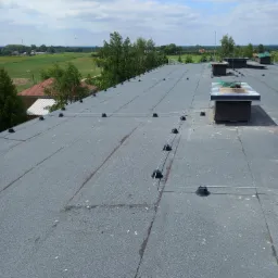 Szary płaski dach pokryty papą z systemem odgromowym, widok z góry na tle zieleni i błękitnego nieba z chmurami. Widoczne kominy wentylacyjne.