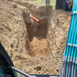 P.H.U. Dr Volt Zbigniew Frejman - Wykop pod instalację sanitarną z widoczną rurą i fragmentem koparki. W tle osoba w ubraniach roboczych i budynek w trakcie budowy.