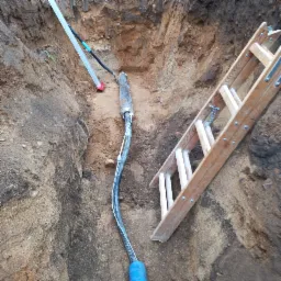 Kabel w wykopie ziemnym, widoczne rury osłonowe, drabina drewniana. Przyłącze kablowe w trakcie realizacji, ziemia piaszczysta, brązowa.