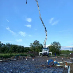 Zbrojenie fundamentów z folią i wystającymi rurami kanalizacyjnymi, na tle pompy do betonu MAN i błękitnego nieba z chmurami.