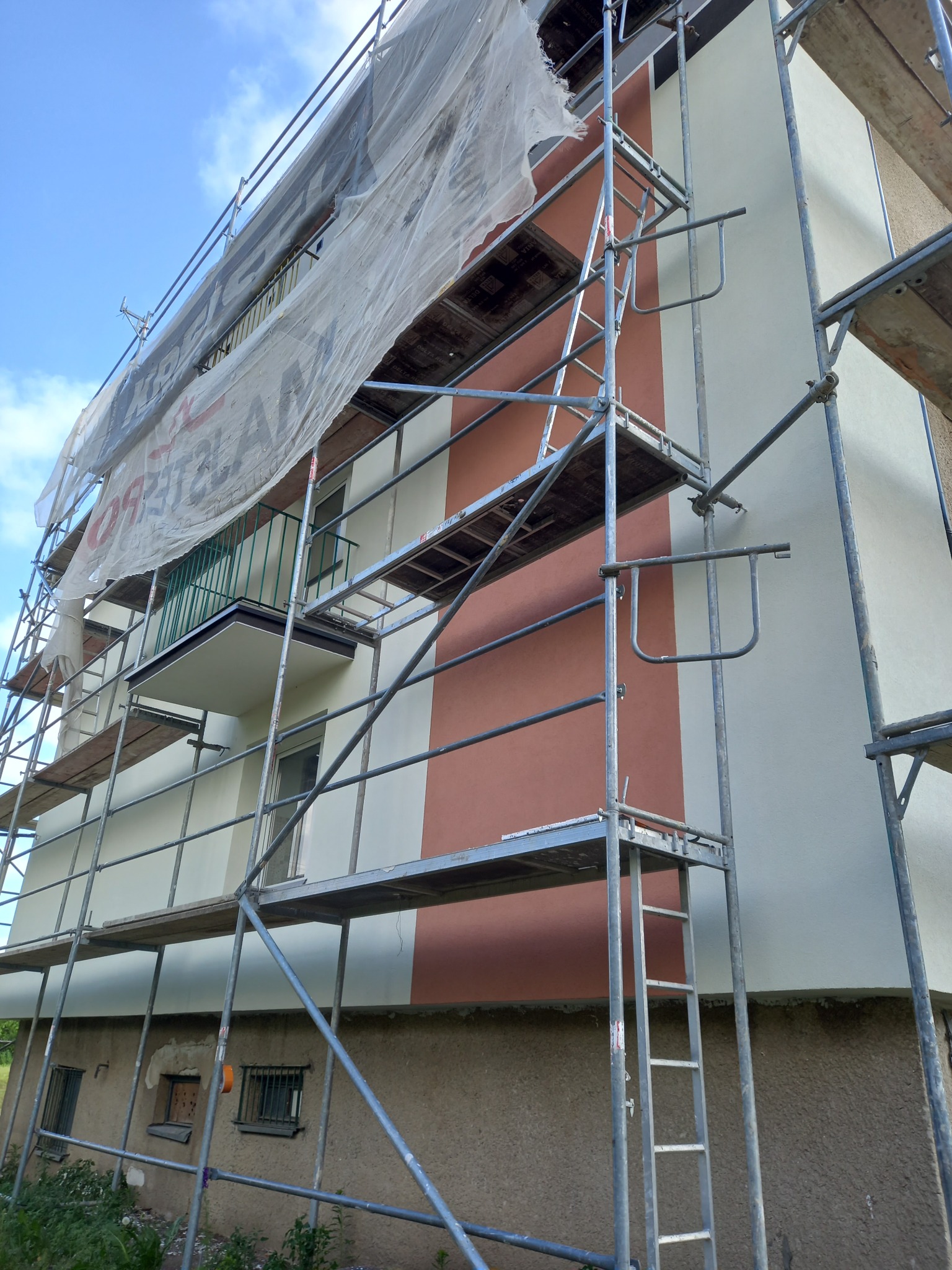 Budynek w rusztowaniach podczas malowania elewacji w pasy, widok z dołu na balkon i fasadę. Prace remontowe w toku, osłonięte siatką ochronną.