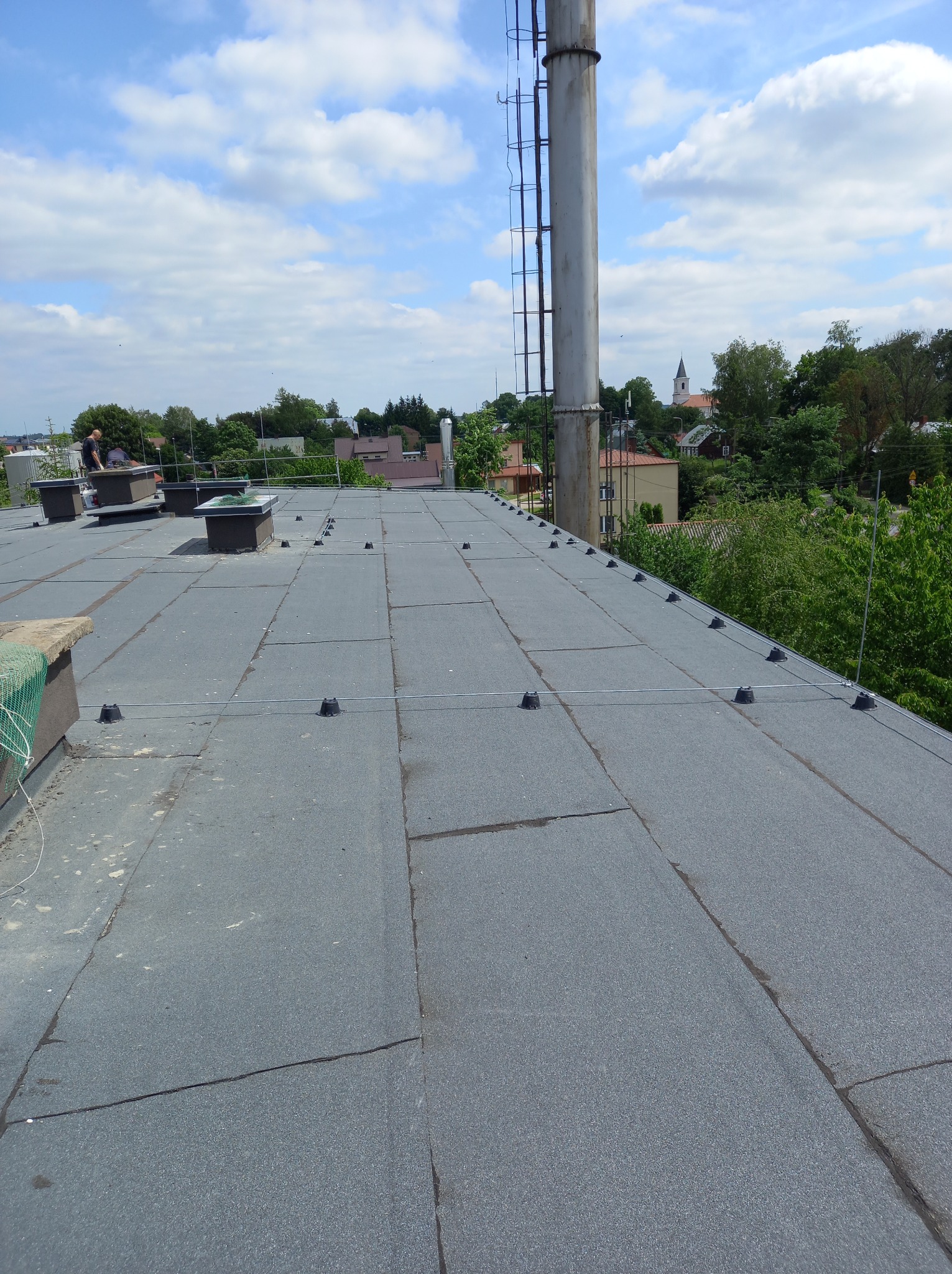 Szary, płaski dach pokryty papą z elementami instalacji odgromowej i kominami wentylacyjnymi. W tle zabudowa miejska i błękitne niebo z chmurami.