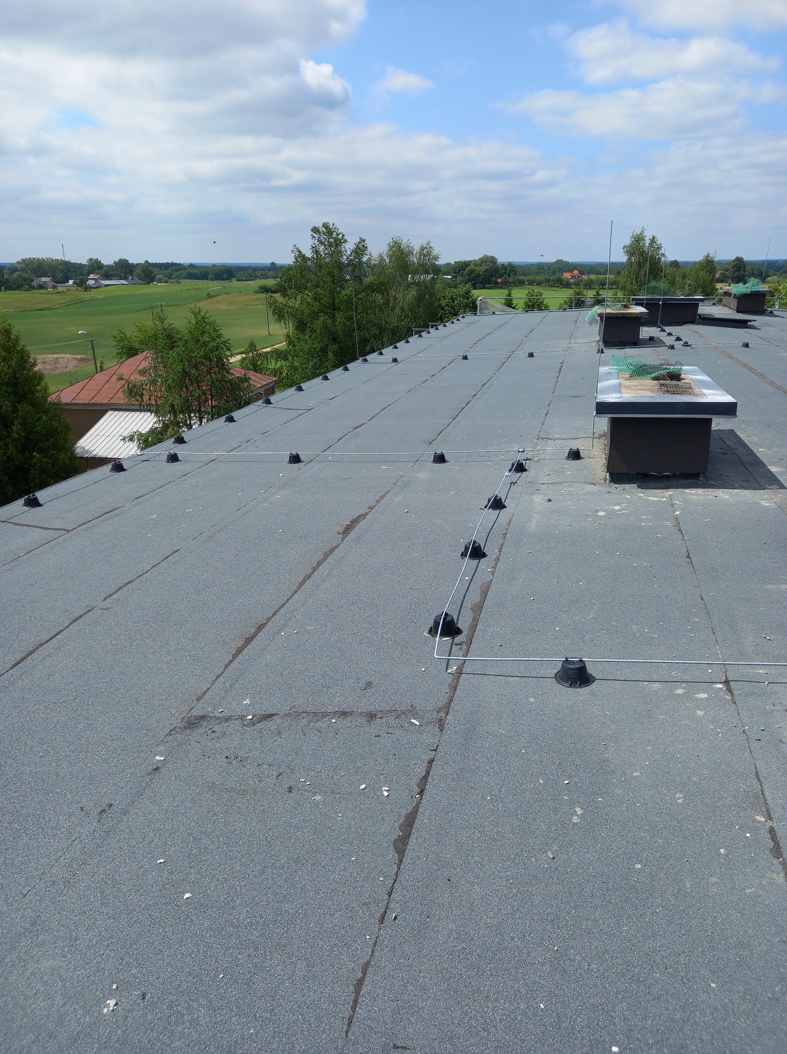 Szary płaski dach pokryty papą z systemem odgromowym, widok z góry na tle zieleni i błękitnego nieba z chmurami. Widoczne kominy wentylacyjne.