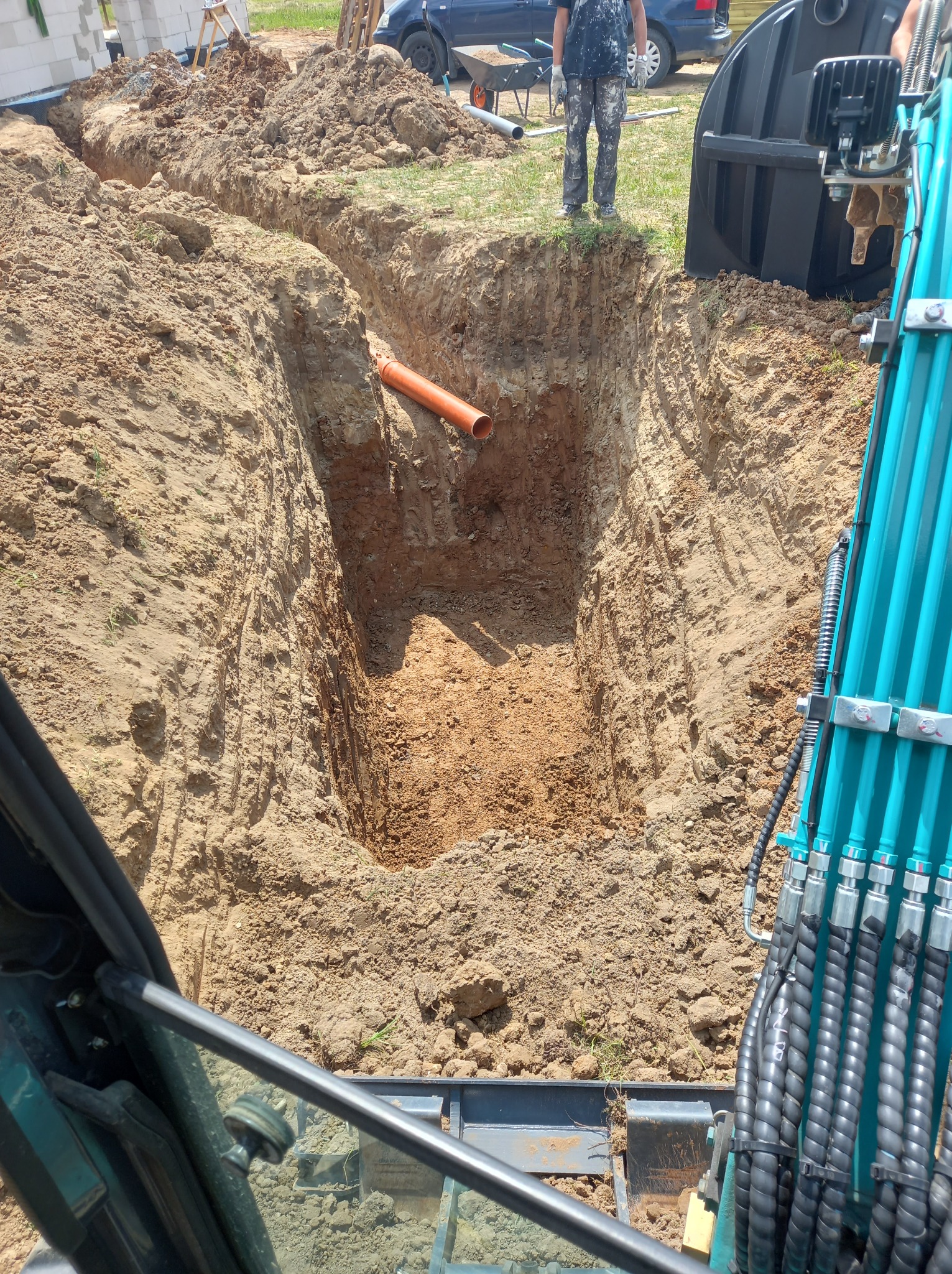 Wykop pod instalację sanitarną z widoczną rurą i fragmentem koparki. W tle osoba w ubraniach roboczych i budynek w trakcie budowy.
