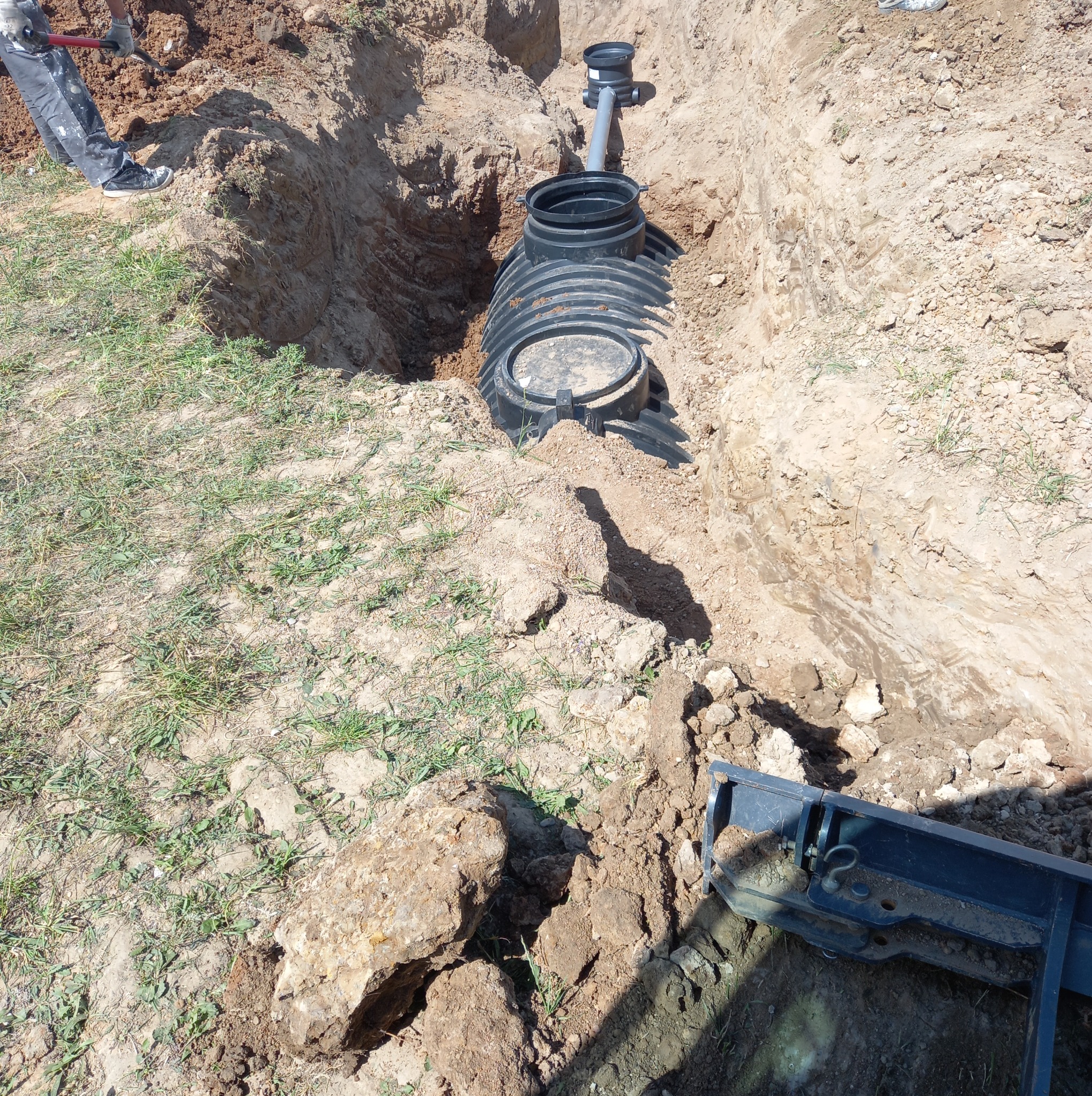 Montaż czarnego, plastikowego zbiornika na deszczówkę w wykopie. Widoczny fragment rury odpływowej i pracownik z łopatą. Realizacja przyłącza wod-kan na działce.