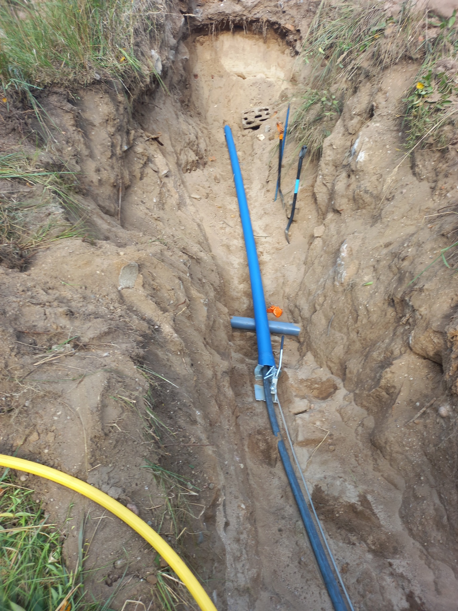 Wykop z ułożonymi rurami (niebieska, szara, żółta) i kablami w ziemi. Widoczne mocowania. Prace ziemne w toku, etap budowy przyłącza.