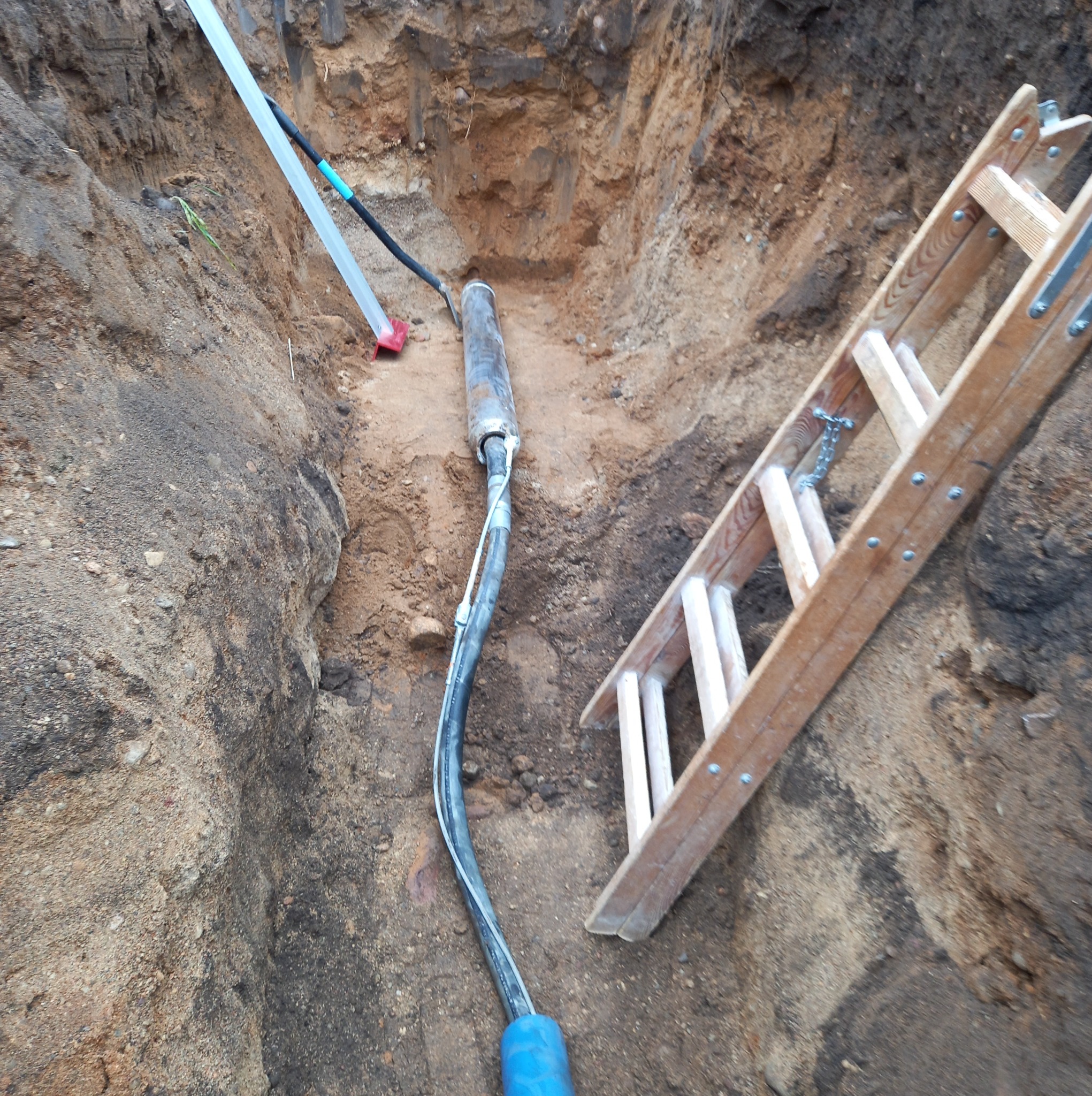 Kabel w wykopie ziemnym, widoczne rury osłonowe, drabina drewniana. Przyłącze kablowe w trakcie realizacji, ziemia piaszczysta, brązowa.